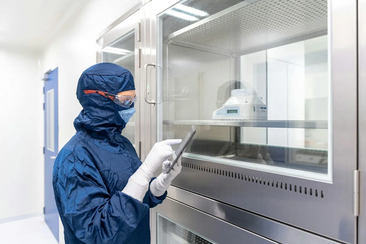 Person in blauer Reinraumkleidung mit Schutzbrille bedient ein Tablet vor einem Edelstahl-Laborschrank
